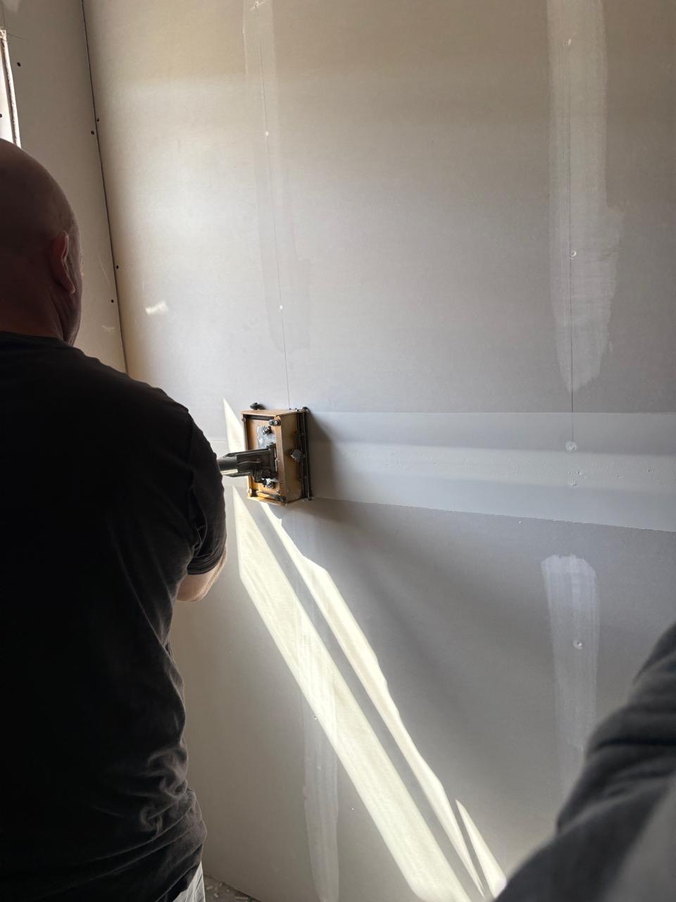 A worker seen from behind runs a flat-box drywall finishing tool along a taped horizontal joint on a large plasterboard wall