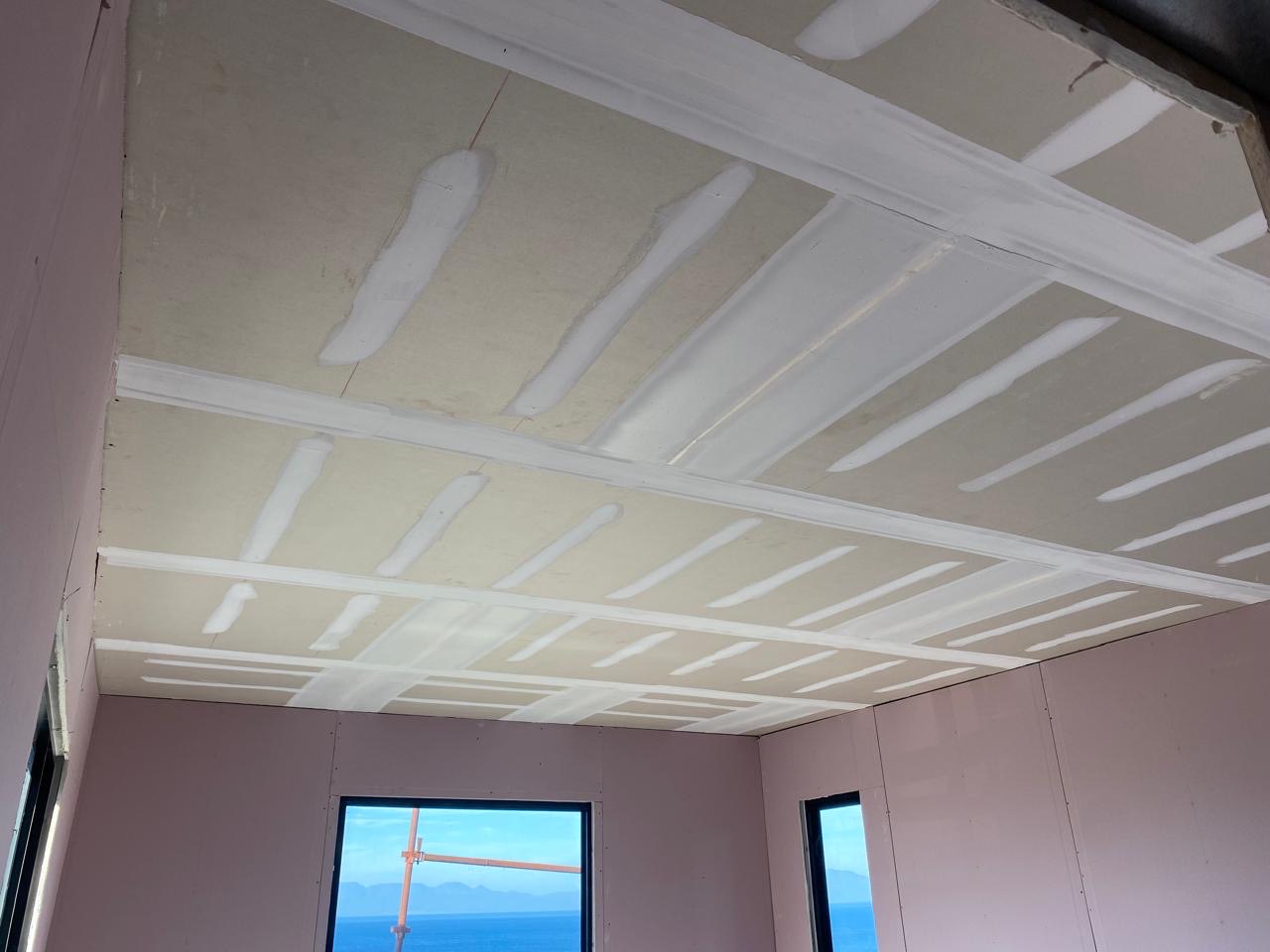 Interior room with drywall ceiling and walls at the taping stage: joints and screw heads coated in white compound in neat bands across the boards