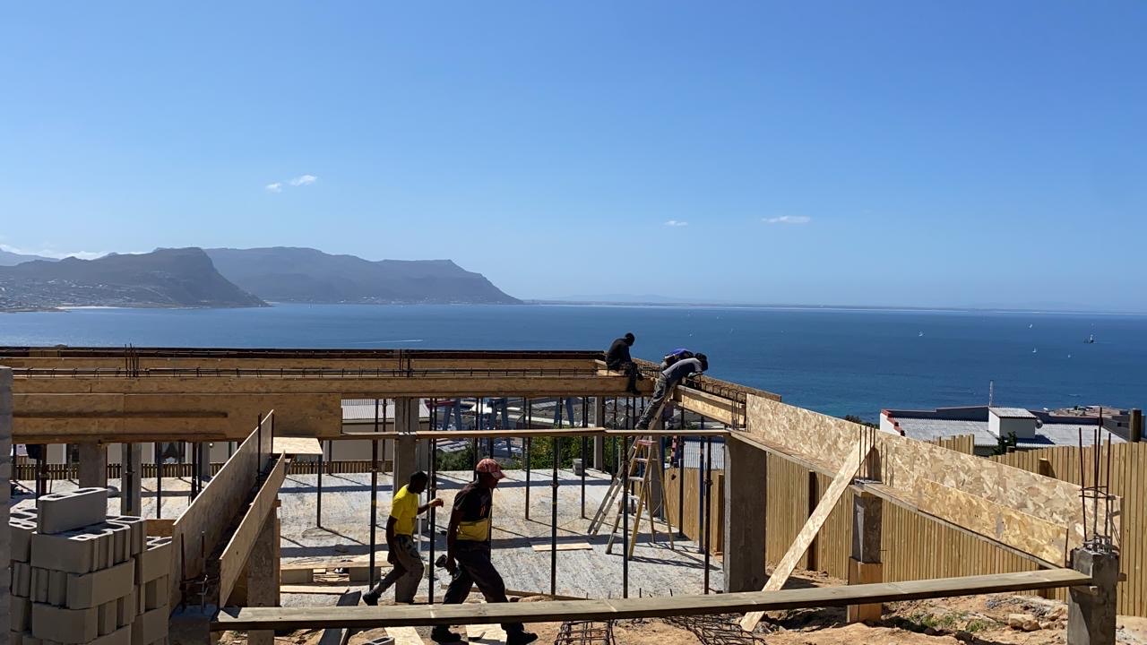 Concrete stage of a hybrid build on a slope above False Bay: shuttered beams with exposed rebar, concrete columns, board formwork and stacked blocks, with workers on the deck and walking through the site
