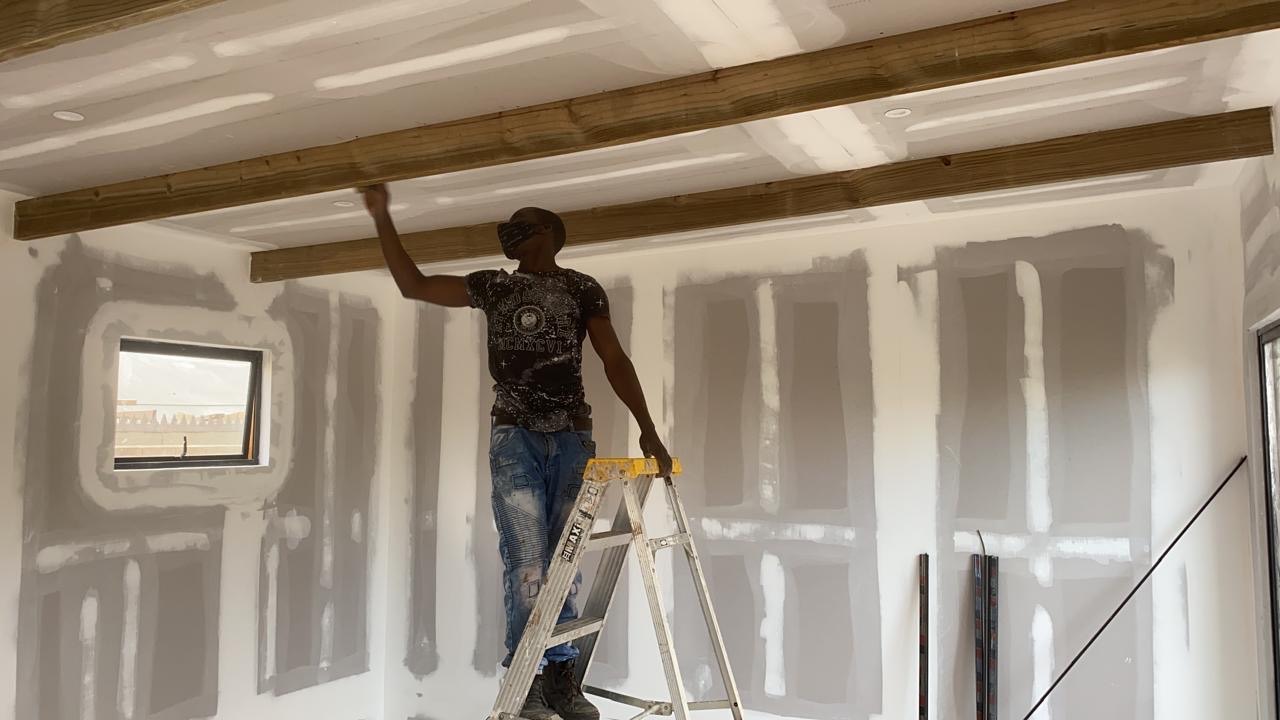 A worker on a stepladder positioning an exposed timber ceiling beam beneath a taped drywall ceiling, with taped and mudded drywall walls behind him