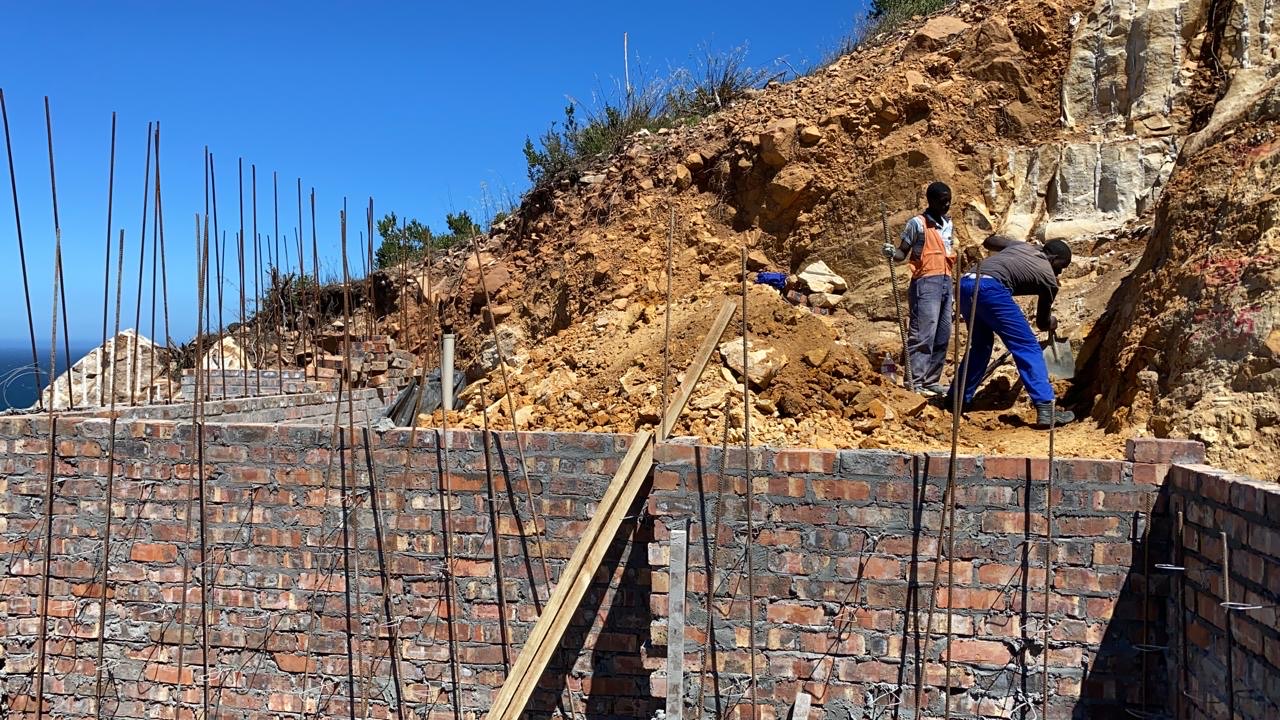 Brick foundation walls with rebar starter bars sticking up, built into a steep rocky excavation with the sea visible at the edge of frame