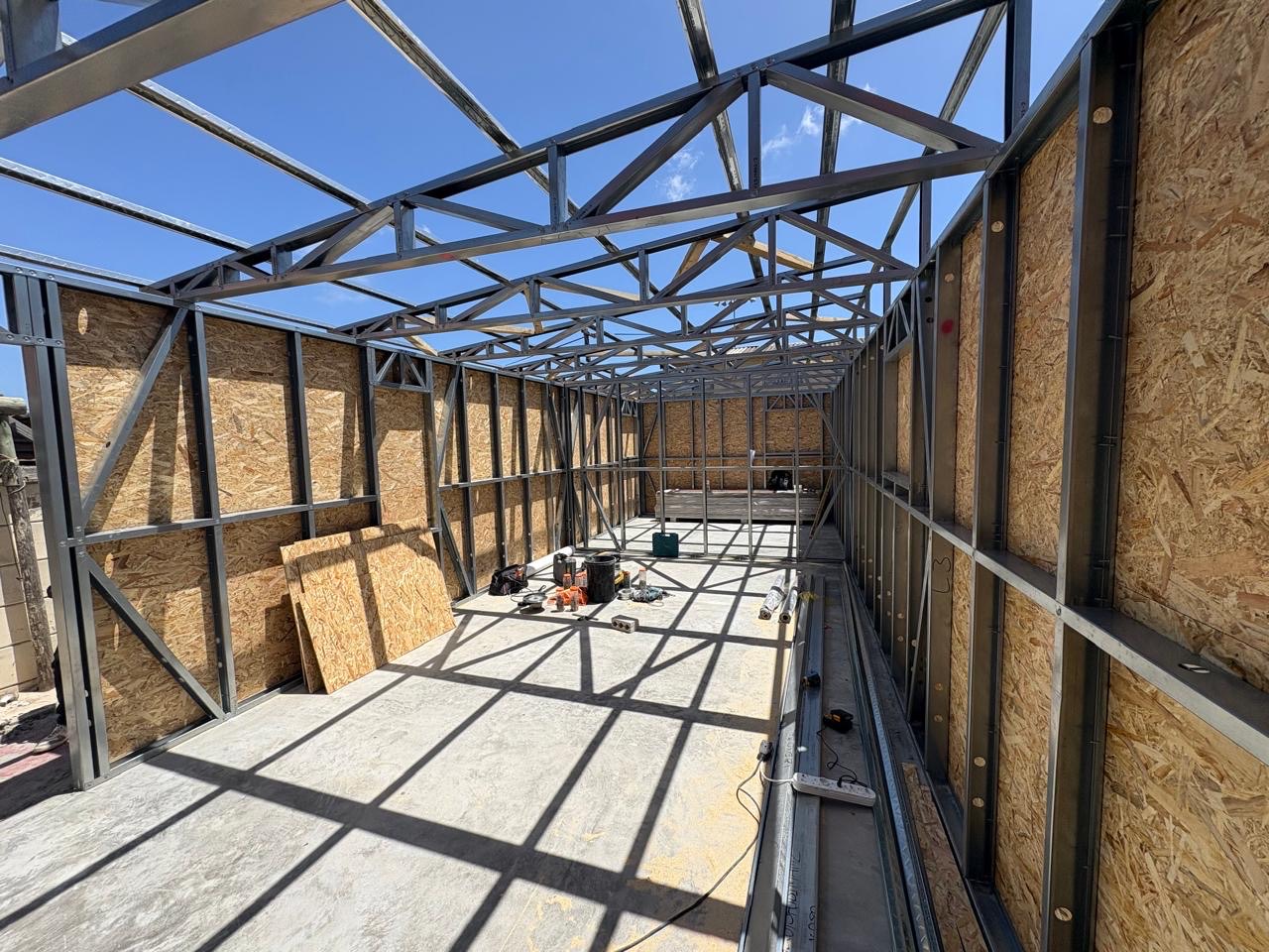 Interior view of a light steel frame structure on a concrete slab, with galvanized wall studs, steel roof trusses open to the sky, and board sheathing fixed to the outside of the walls