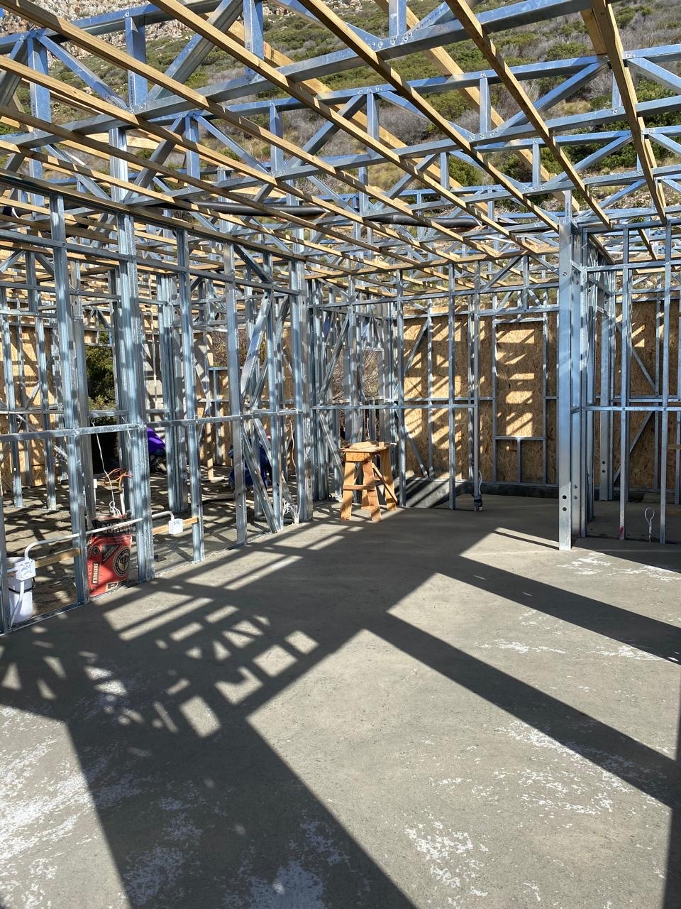 Inside a light steel frame structure on a concrete slab, with galvanised wall studs, steel roof trusses and timber purlins overhead, and OSB sheathing on the far walls