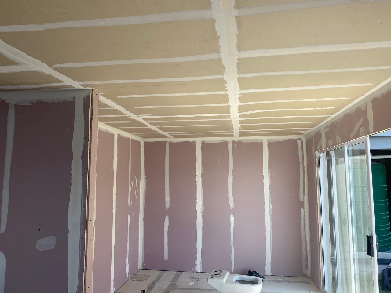 Interior room with pink/grey drywall boards on walls and ceiling, all joints taped and mudded but not yet skimmed
