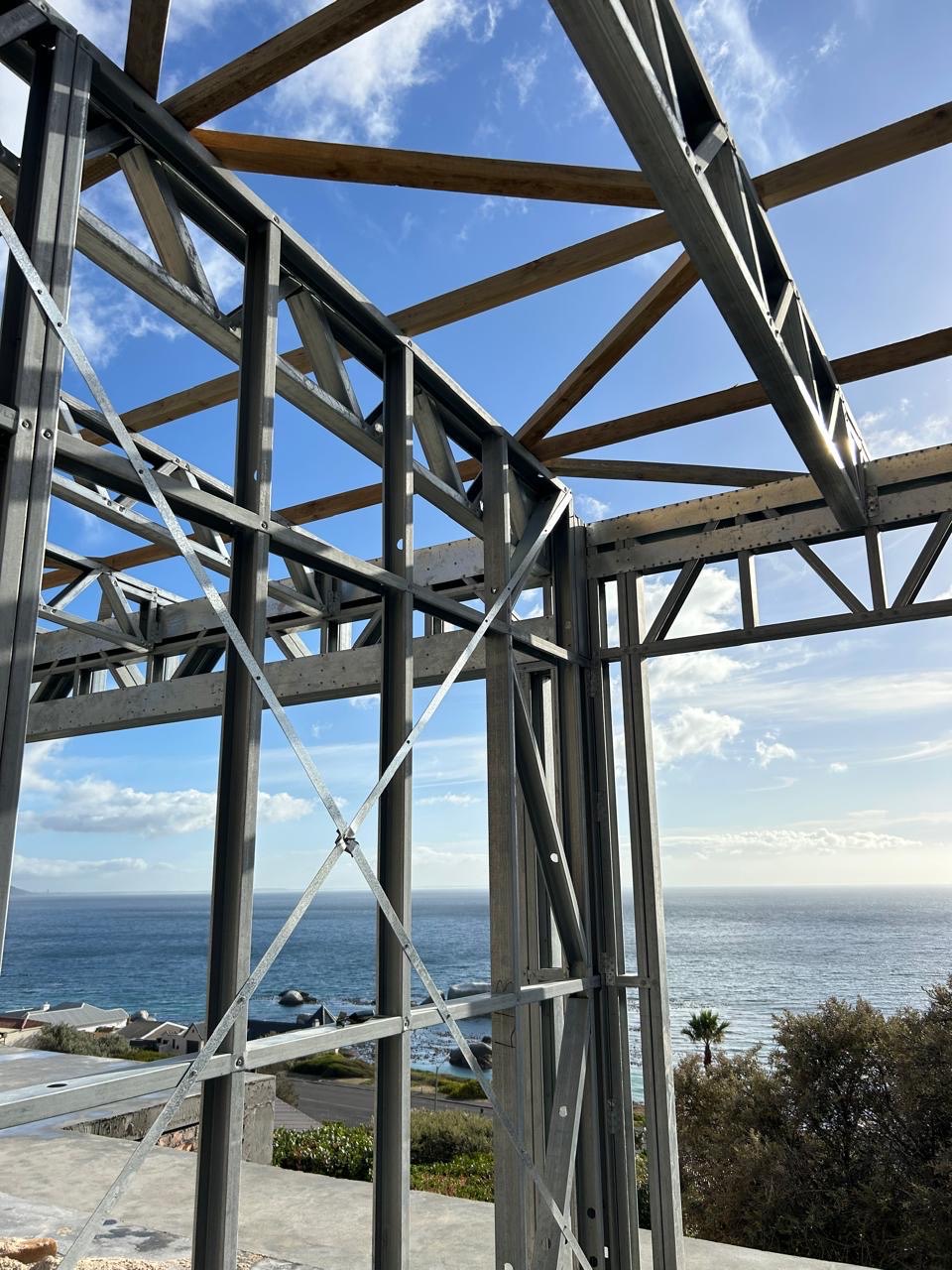 Looking up through a light-steel frame with timber roof battens being fixed over the truss beams, with a coastal neighbourhood, granite boulders and the sea below