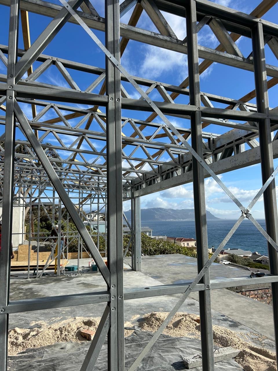 Looking through a Zincalume steel frame from inside a build, with lattice roof trusses and timber battens overhead and the concrete slab below