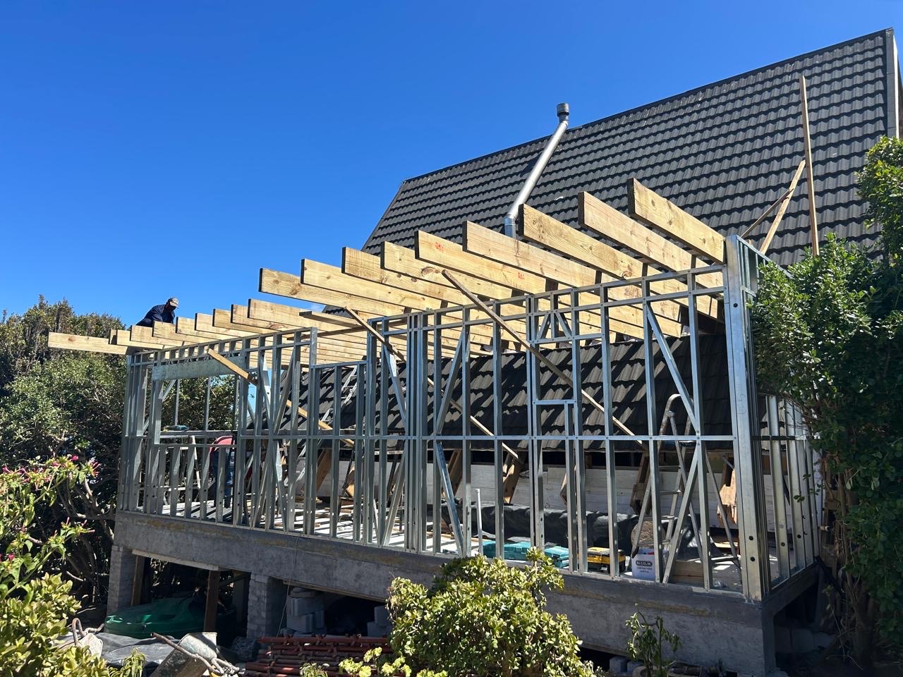 A first-floor extension being added to an existing tiled-roof house: light-steel wall frames stand on a masonry base with timber joists laid across the top