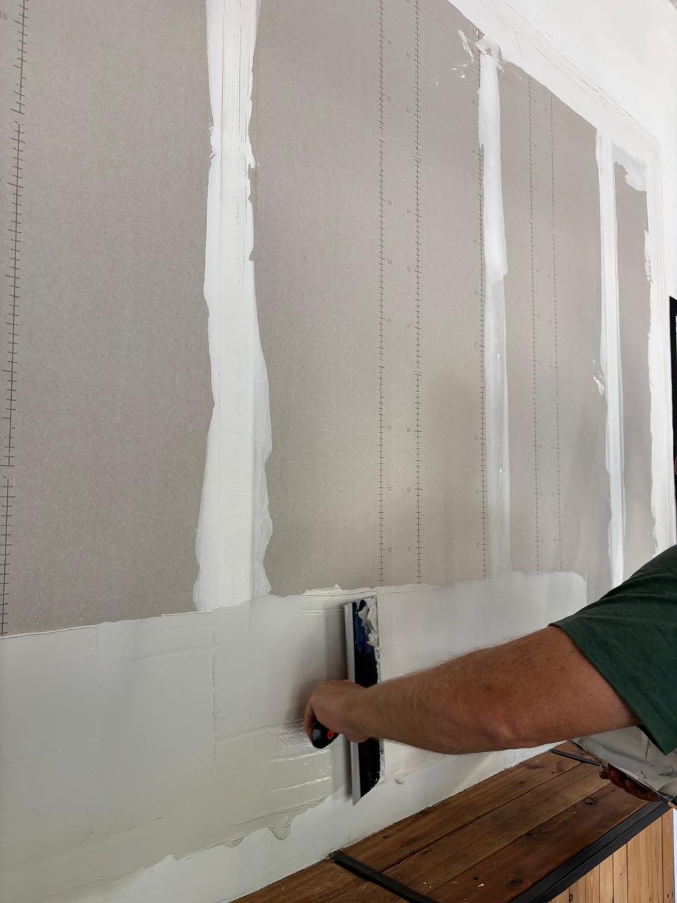 A close-up of someone applying a skim coat to a taped drywall wall with a wide finishing blade