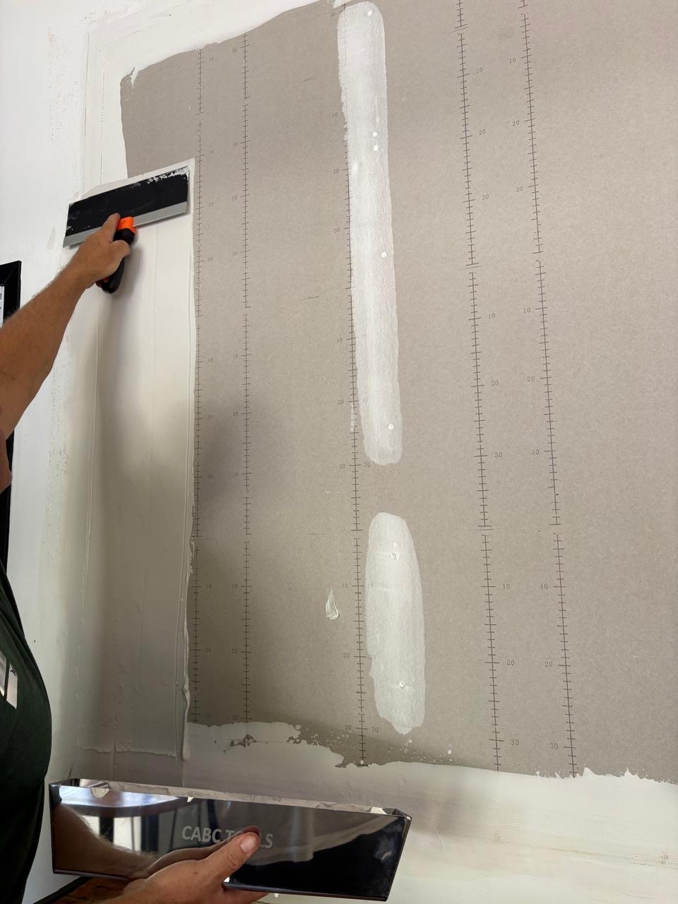 Close-up of someone pulling a skim coat over drywall with a wide blade, one taped joint already coated smooth, while their other hand holds a stainless mud pan marked CABC TOOLS
