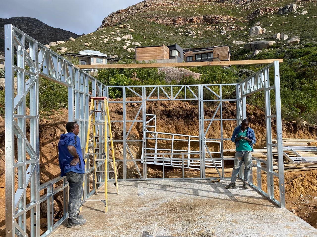 Two workers erecting galvanized light steel wall frames on a concrete slab, with a yellow ladder between the panels and a rocky Cape Town hillside with modern houses behind them
