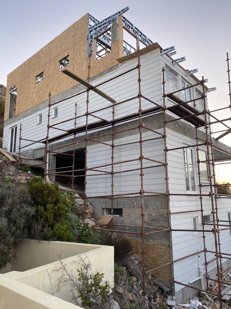 A multi-storey house on a rocky slope wrapped in steel scaffolding, with white horizontal cladding and a concrete band on the lower floors and a board-sheathed light steel frame forming the top storey