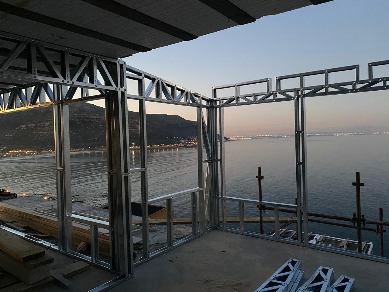 Zincalume light-steel wall frames and lattice trusses on an upper floor at dusk, shot from inside the structure looking out over a bay with mountains and town lights in the distance