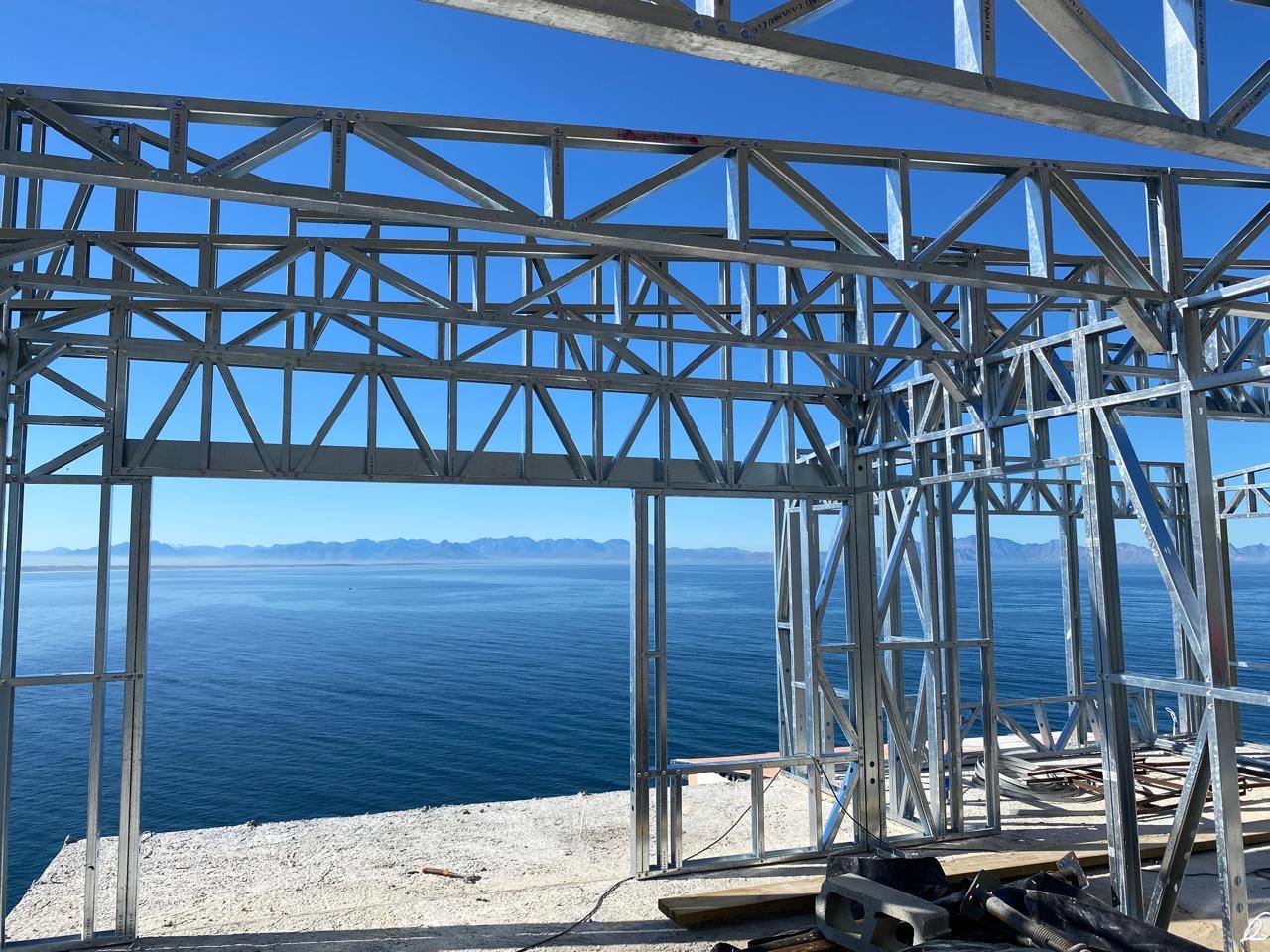 View from inside an erected light-steel frame, with lattice truss beams and stud walls framing a wide ocean vista and distant mountains under a clear blue sky