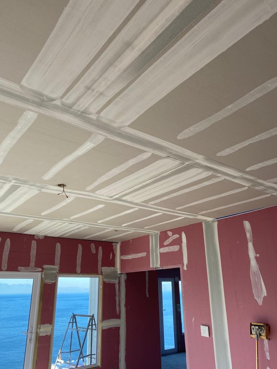 Room interior mid-finishing: the plasterboard ceiling has taped and coated joints and screw lines, while the pink fire-rated boards on the walls show patched fixings