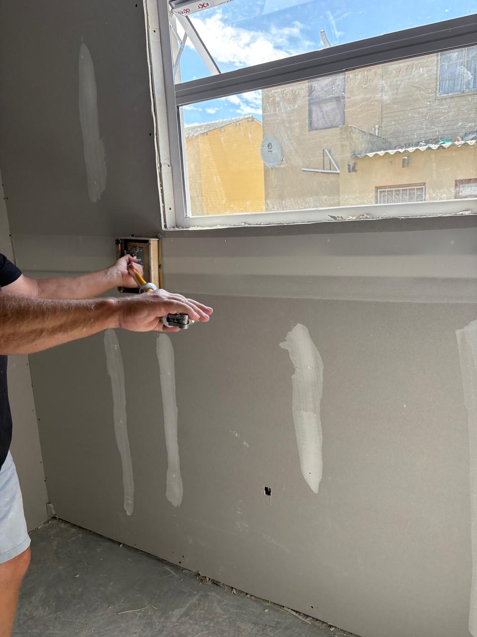 A worker's arms applying joint compound to drywall below a window, with taped and coated joints visible on the boarded walls; only hands and forearms are in frame