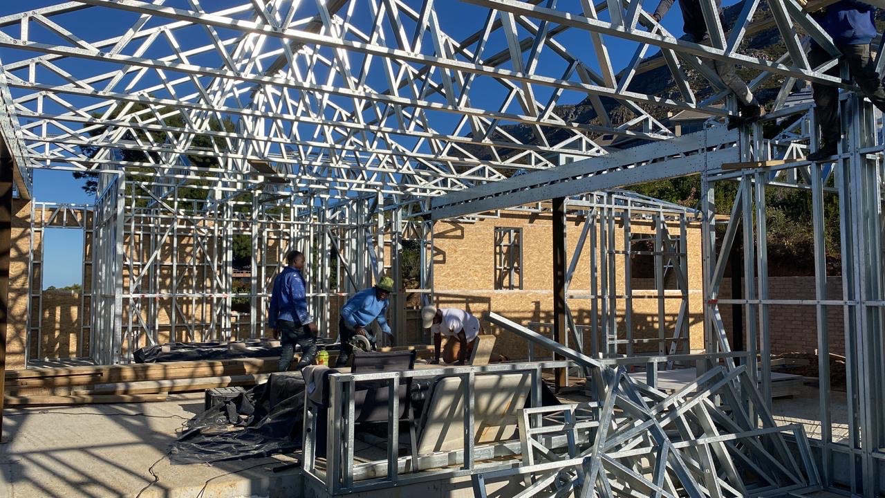 Crew working inside a large light-steel frame with trusses already up overhead, board sheathing fixed to some wall panels in the background and workers climbing the frame at top right