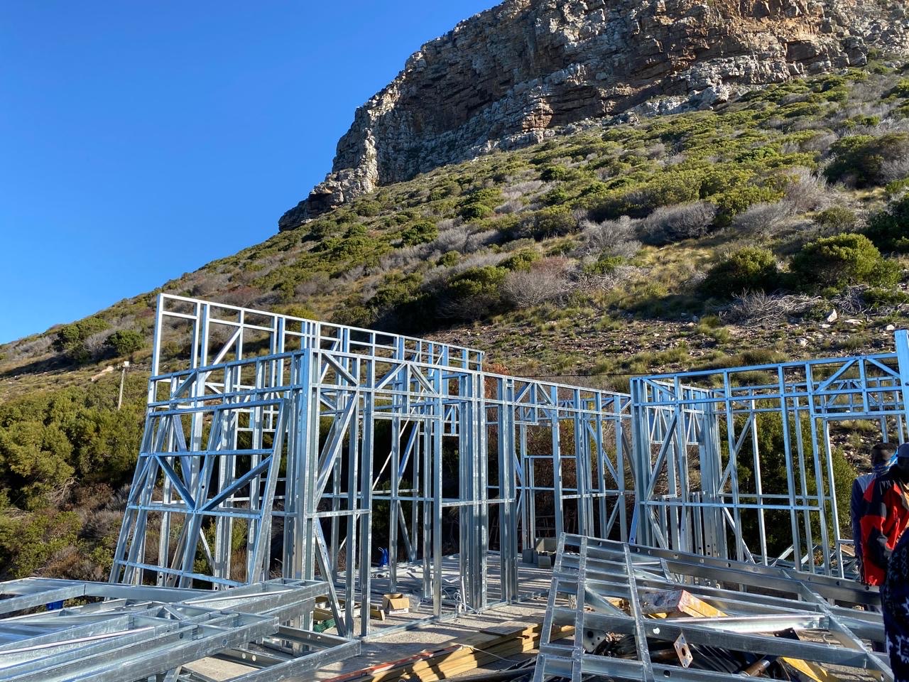 Light-steel wall frames erected on a slab at the foot of a rocky, fynbos-covered mountainside, with prefabricated steel trusses and timber stacked in the foreground