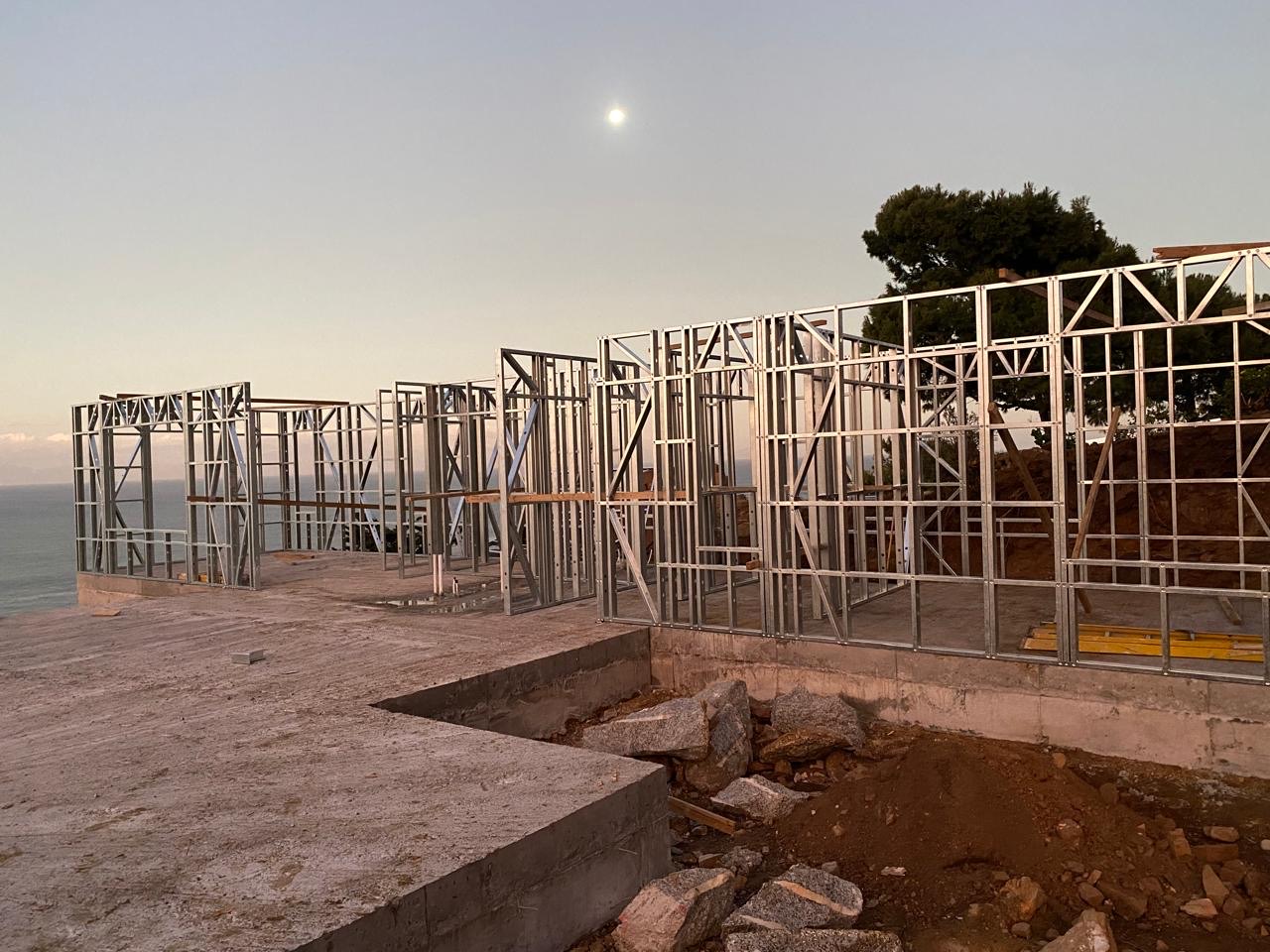 Galvanized light-steel wall frame panels standing on a fresh concrete slab at dusk, with the moon overhead, the sea in the background and excavated earth and rock in the foreground